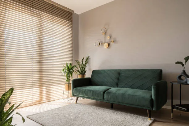Elegant living room with big window wall behind wooden blinds, comfortable green sofa, carpet and decorative gold wall lamp